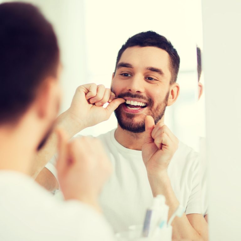 health care, dental hygiene, people and beauty concept - smiling young man with floss cleaning teeth and looking to mirror at home bathroom